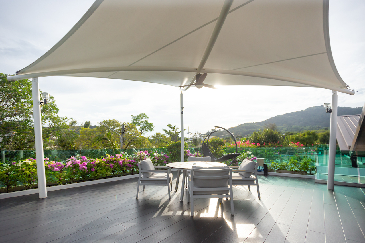 Outdoor terrace with a large curved white shade, round table and four chairs, flowering garden and hills in the background.