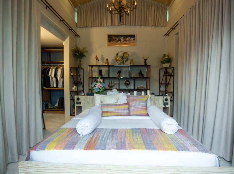 Showroom bedroom with a bed, colorful quilt, and two white bolster pillows centered against a shelving display behind.