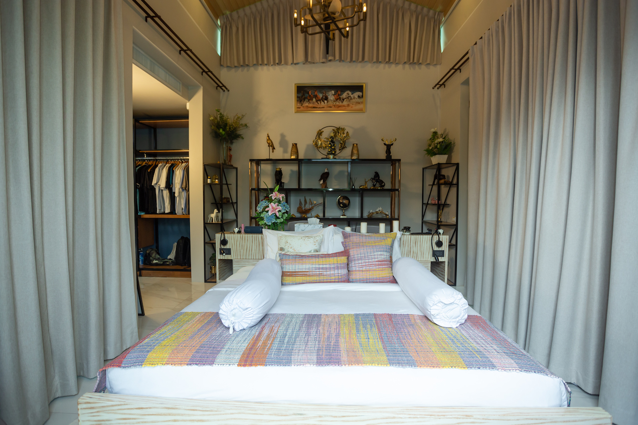 Showroom bedroom with a bed, colorful quilt, and two white bolster pillows centered against a shelving display behind.