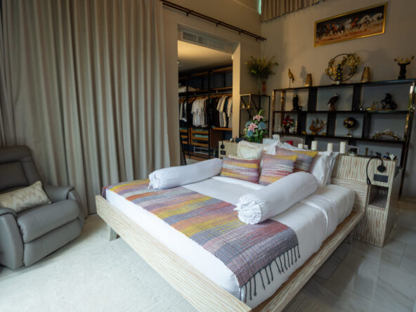 Bedroom with a low wooden platform bed, colorful throw, and rolled white bolster pillows in front of a display shelf.