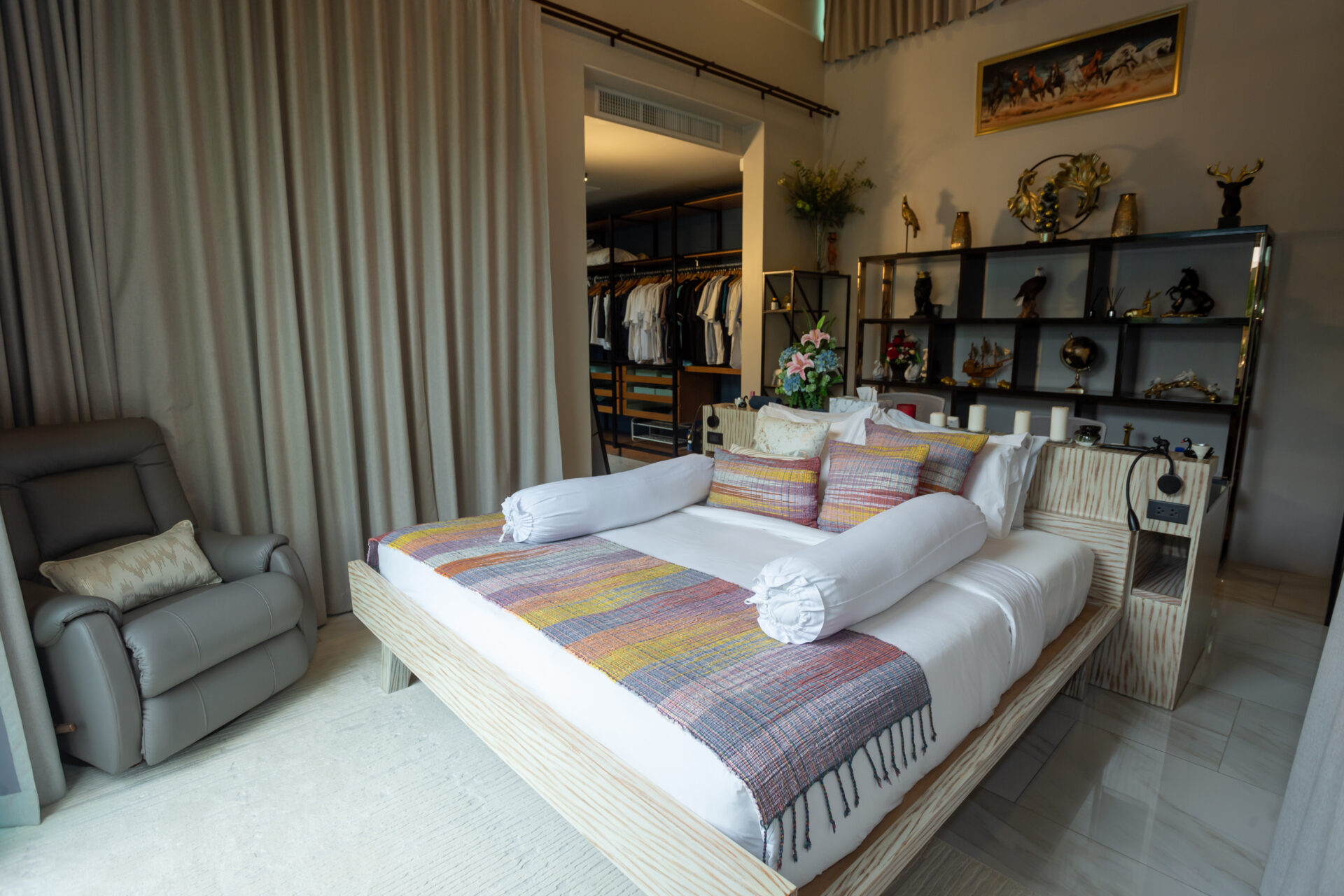 Bedroom with a low wooden platform bed, colorful throw, and rolled white bolster pillows in front of a display shelf.