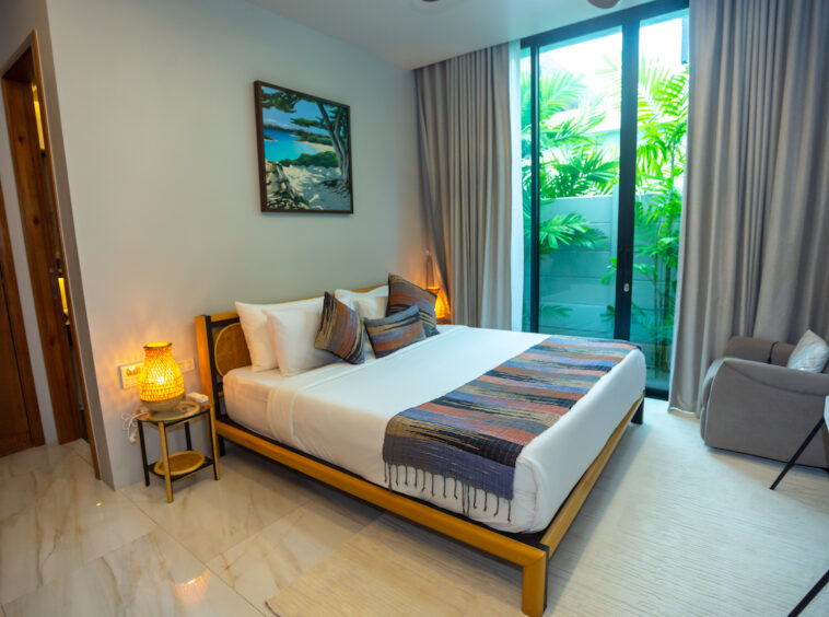 Hotel bedroom with a large bed, warm lamps, and a balcony door revealing tropical plants outside,