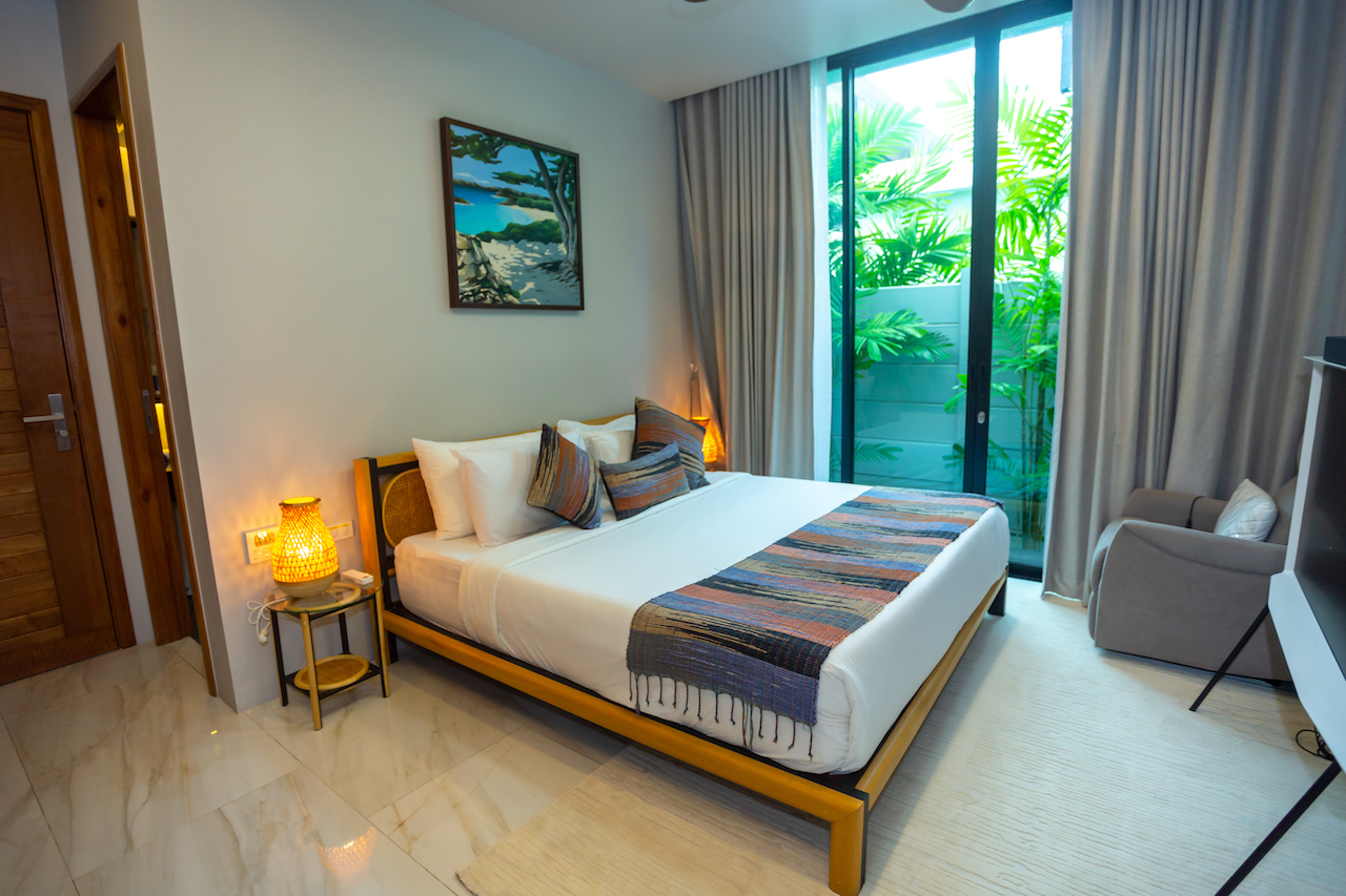 Hotel bedroom with a large bed, warm lamps, and a balcony door revealing tropical plants outside,
