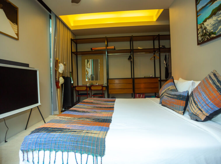 Hotel bedroom with white bed, colorful plaid throws, and an open clothing wardrobe against the back wall under a warm yellow ceiling light.