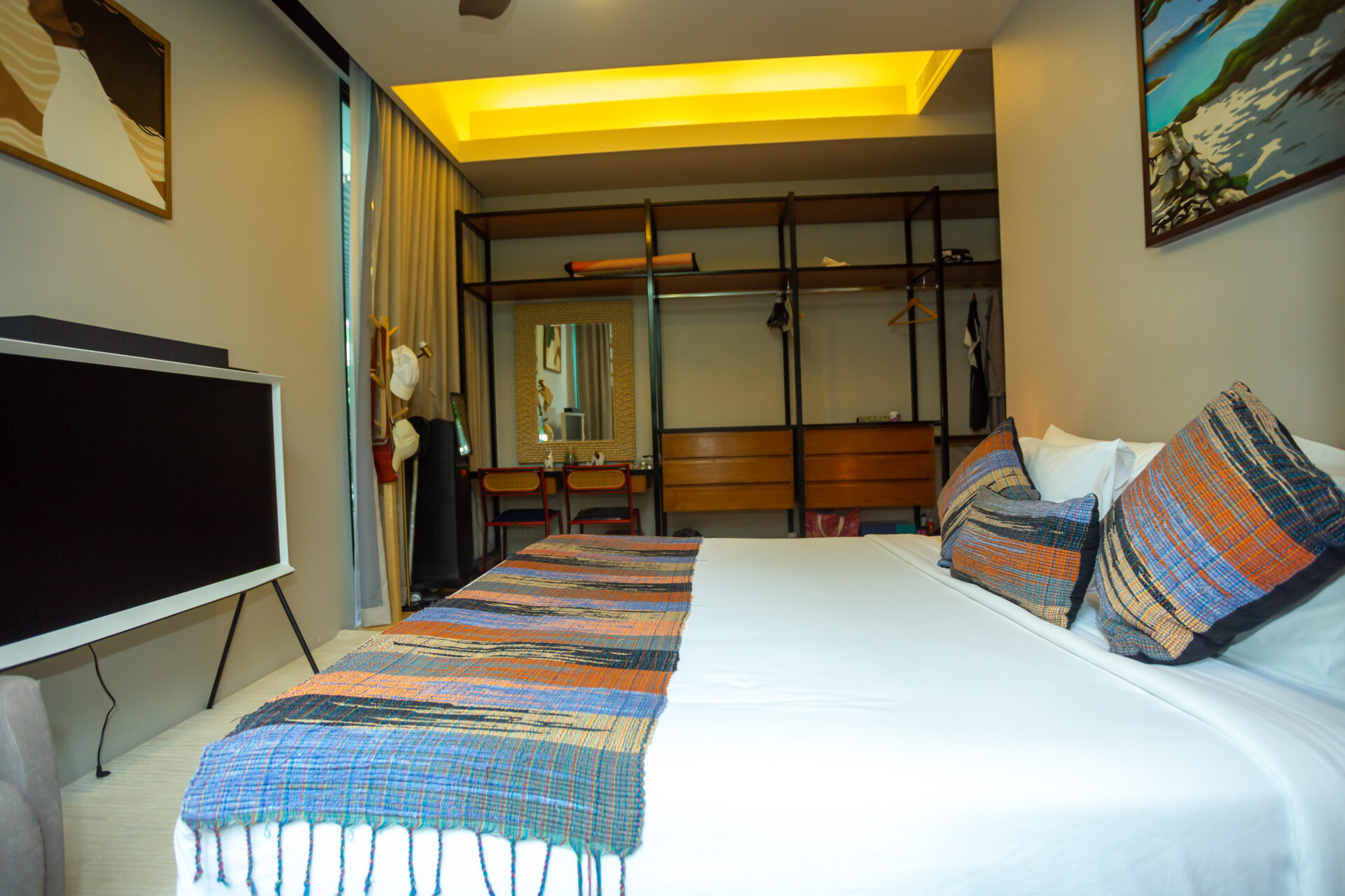 Hotel bedroom with white bed, colorful plaid throws, and an open clothing wardrobe against the back wall under a warm yellow ceiling light.