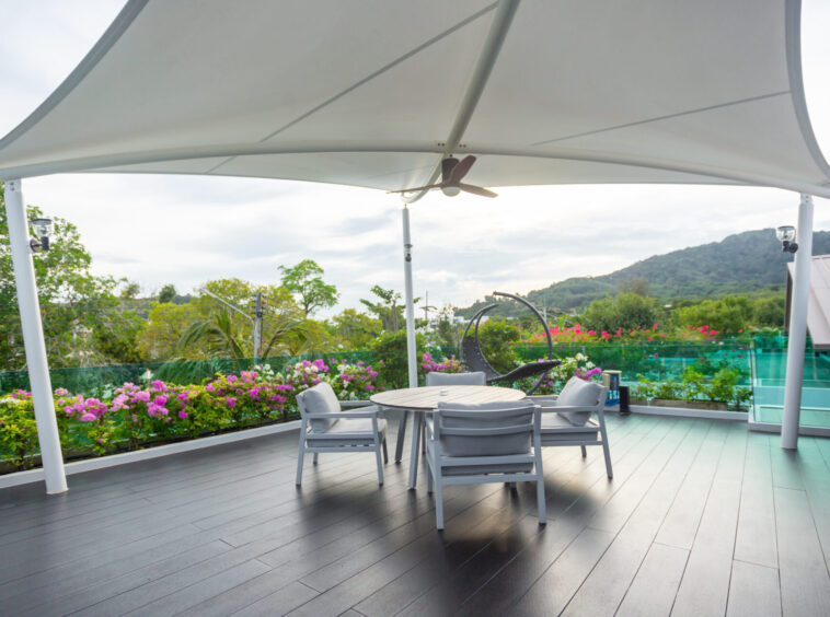 Outdoor patio with a round table and chairs under a large white canopy, lush garden flowers in the background and hills in the distance.