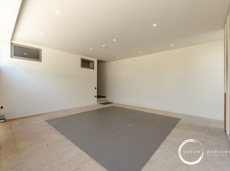 Covered carport with white walls and ceiling, small rectangular windows, and a dark gray tiled area on the floor; doorway to interior on the left and a watermark in the lower right corner.