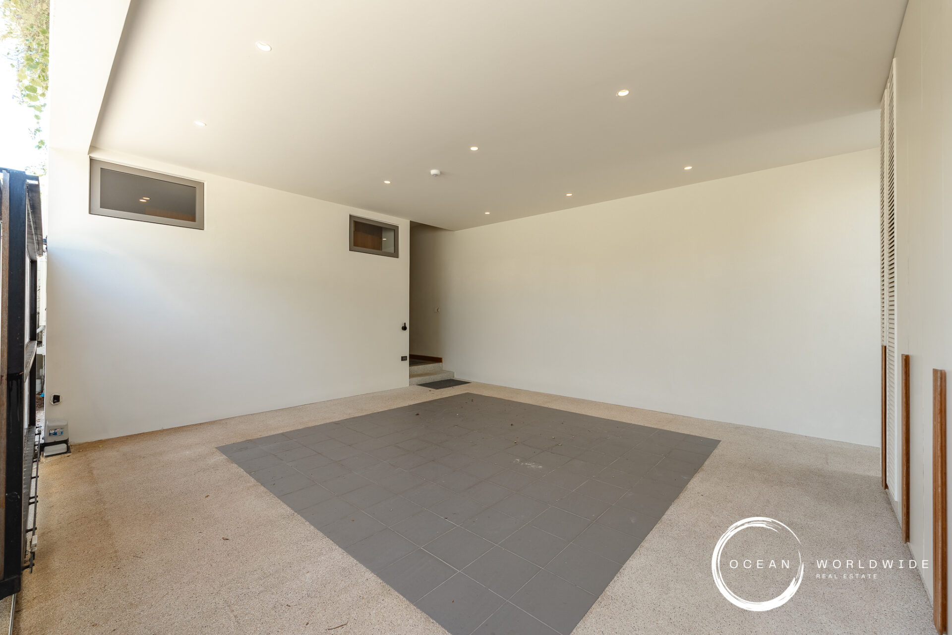 Covered carport with white walls and ceiling, small rectangular windows, and a dark gray tiled area on the floor; doorway to interior on the left and a watermark in the lower right corner.