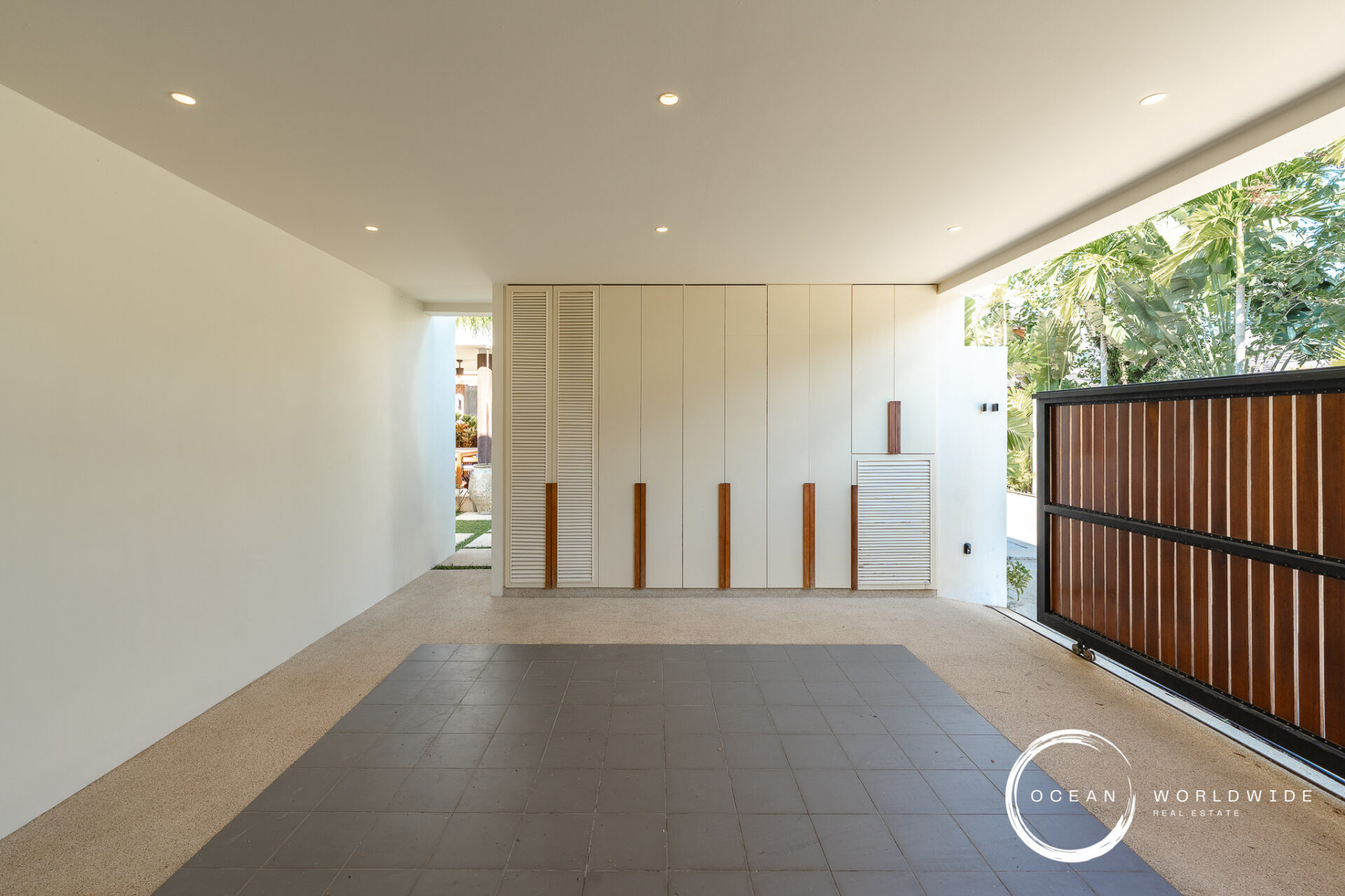 Modern covered outdoor hallway with white storage cabinets and vertical wooden handles, opening to a tropical garden."