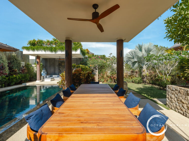 Long wooden outdoor dining table with blue cushions, under a covered patio next to a pool and lush tropical plants.