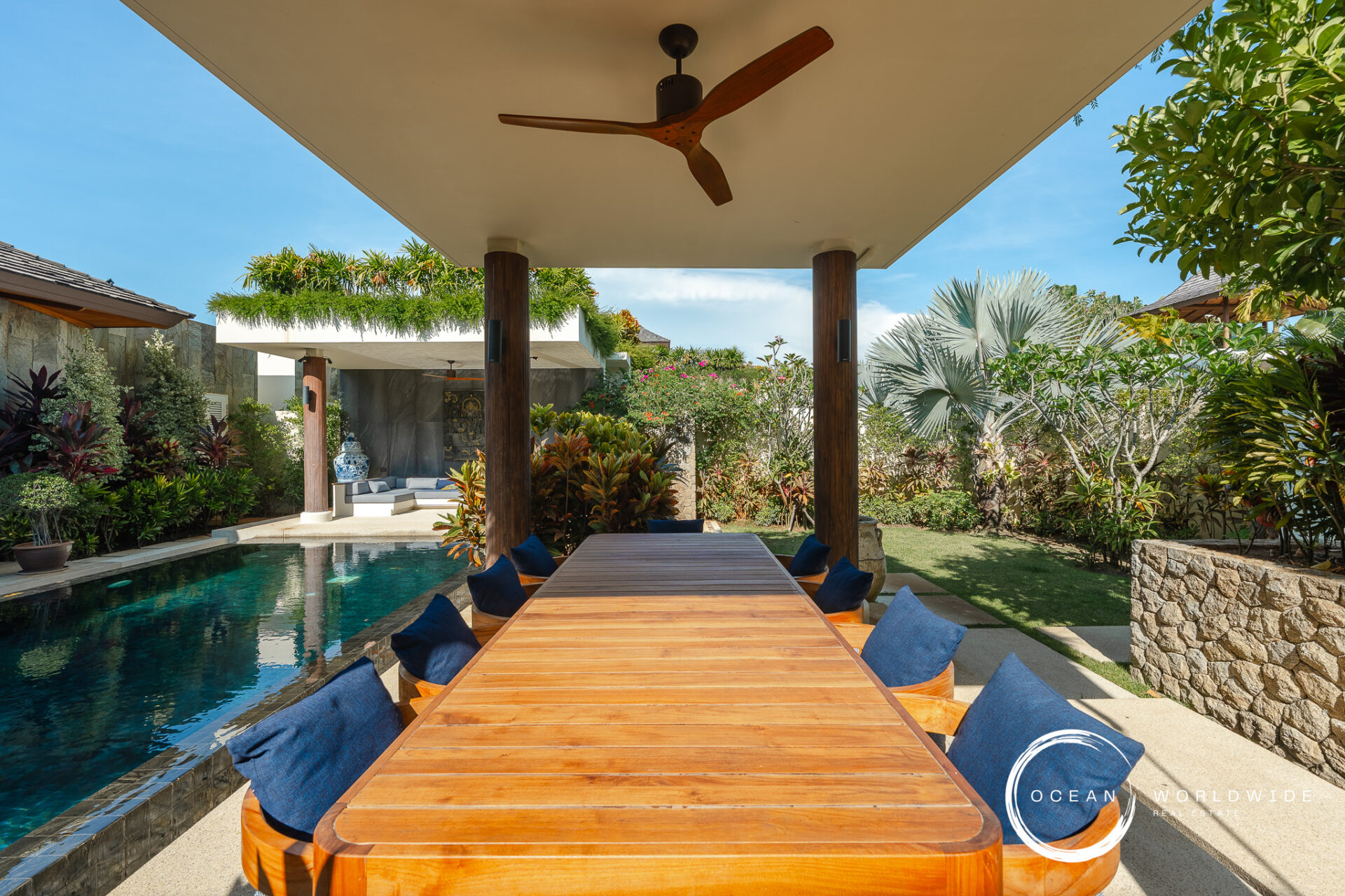 Long wooden outdoor dining table with blue cushions, under a covered patio next to a pool and lush tropical plants.