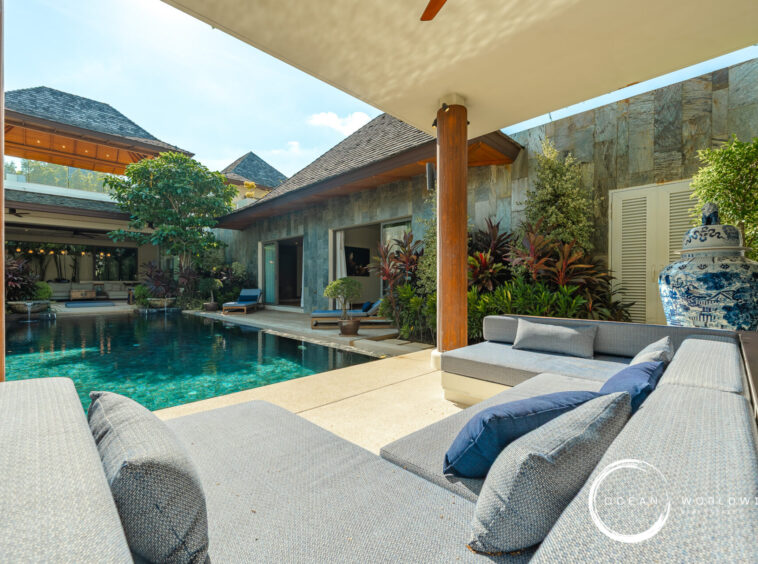 Poolside courtyard of a modern villa with a stone wall, wooden columns, and gray sectional loungers overlooking a turquoise pool.
