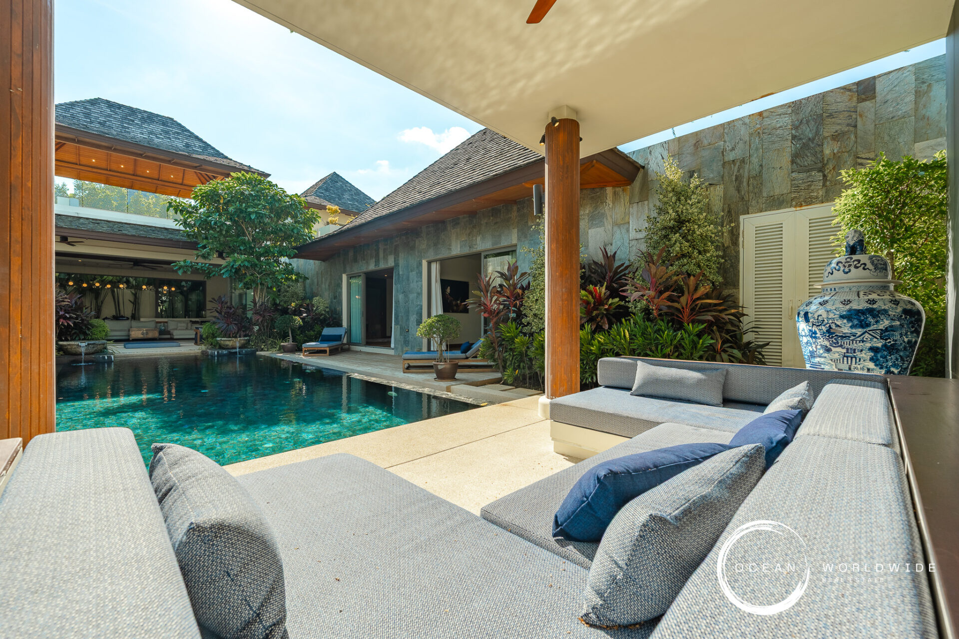Poolside courtyard of a modern villa with a stone wall, wooden columns, and gray sectional loungers overlooking a turquoise pool.