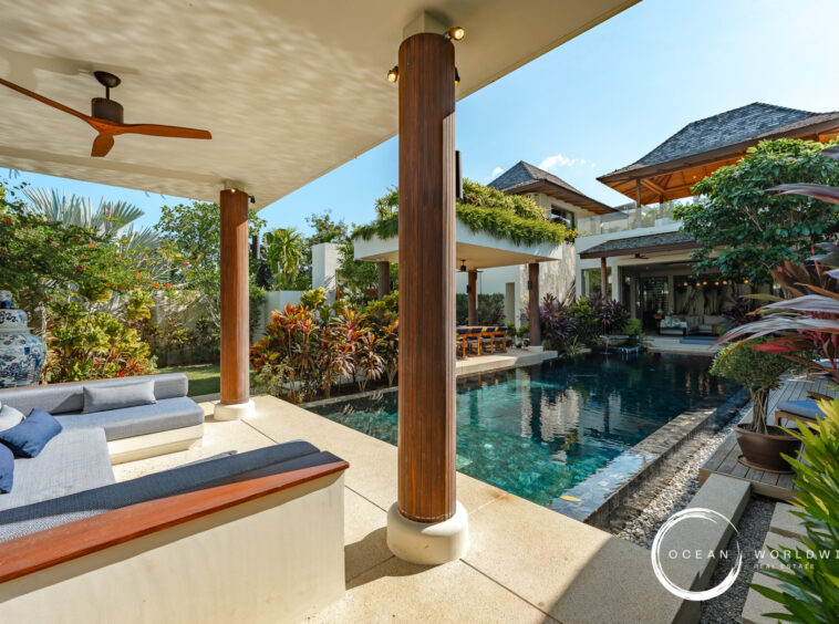Open-air poolside lounge of a luxury villa with wooden columns, a shaded seating area, and tropical plants.