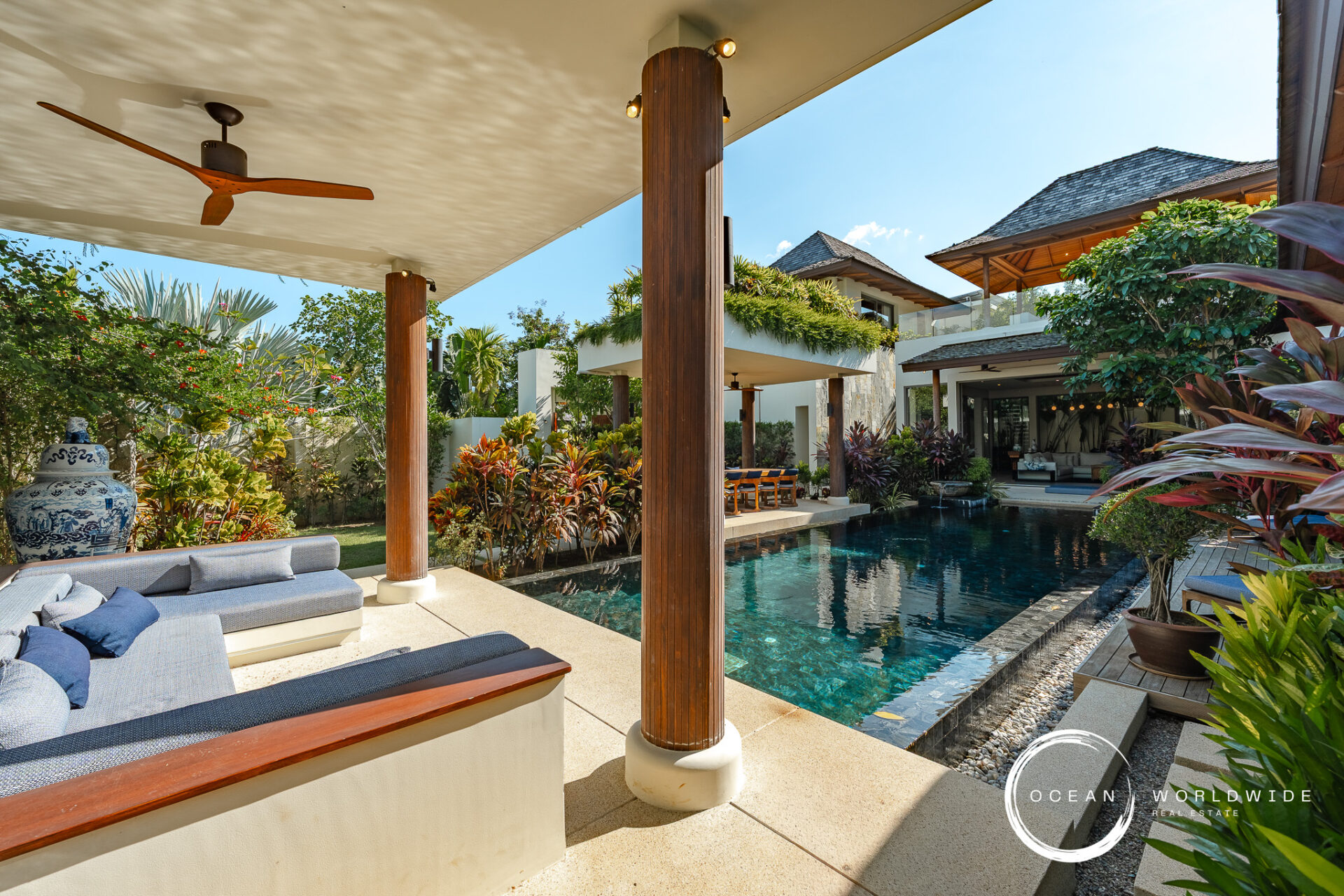 Open-air poolside lounge of a luxury villa with wooden columns, a shaded seating area, and tropical plants.
