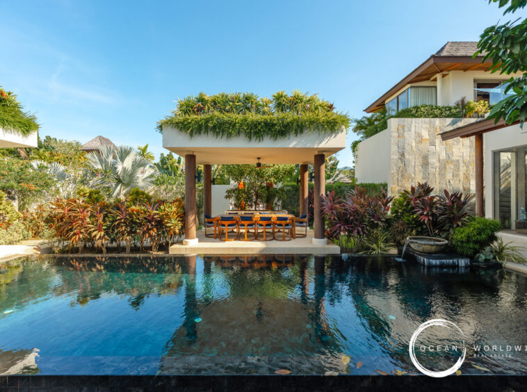 Resort-style backyard with a rectangular pool, a shaded dining area under a pavilion, and tropical plants surrounding a modern villa.