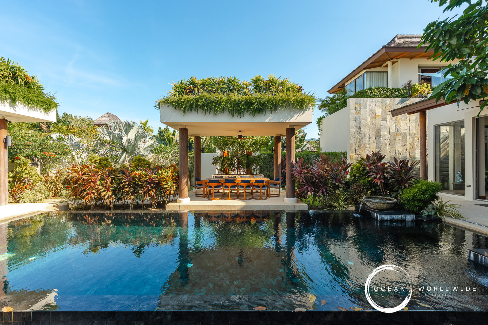 Resort-style backyard with a rectangular pool, a shaded dining area under a pavilion, and tropical plants surrounding a modern villa.