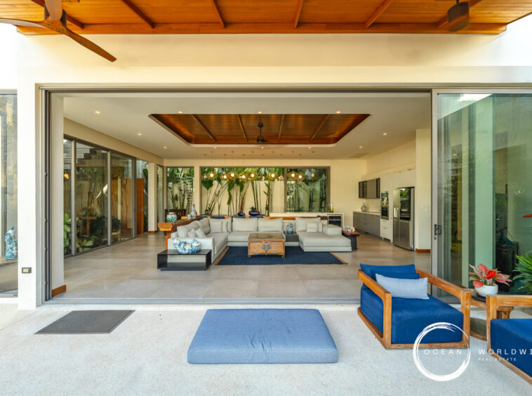 Open-plan living room seen through wide sliding doors; gray sectional sofa, blue cushions, tropical plants, and a kitchen in the background.