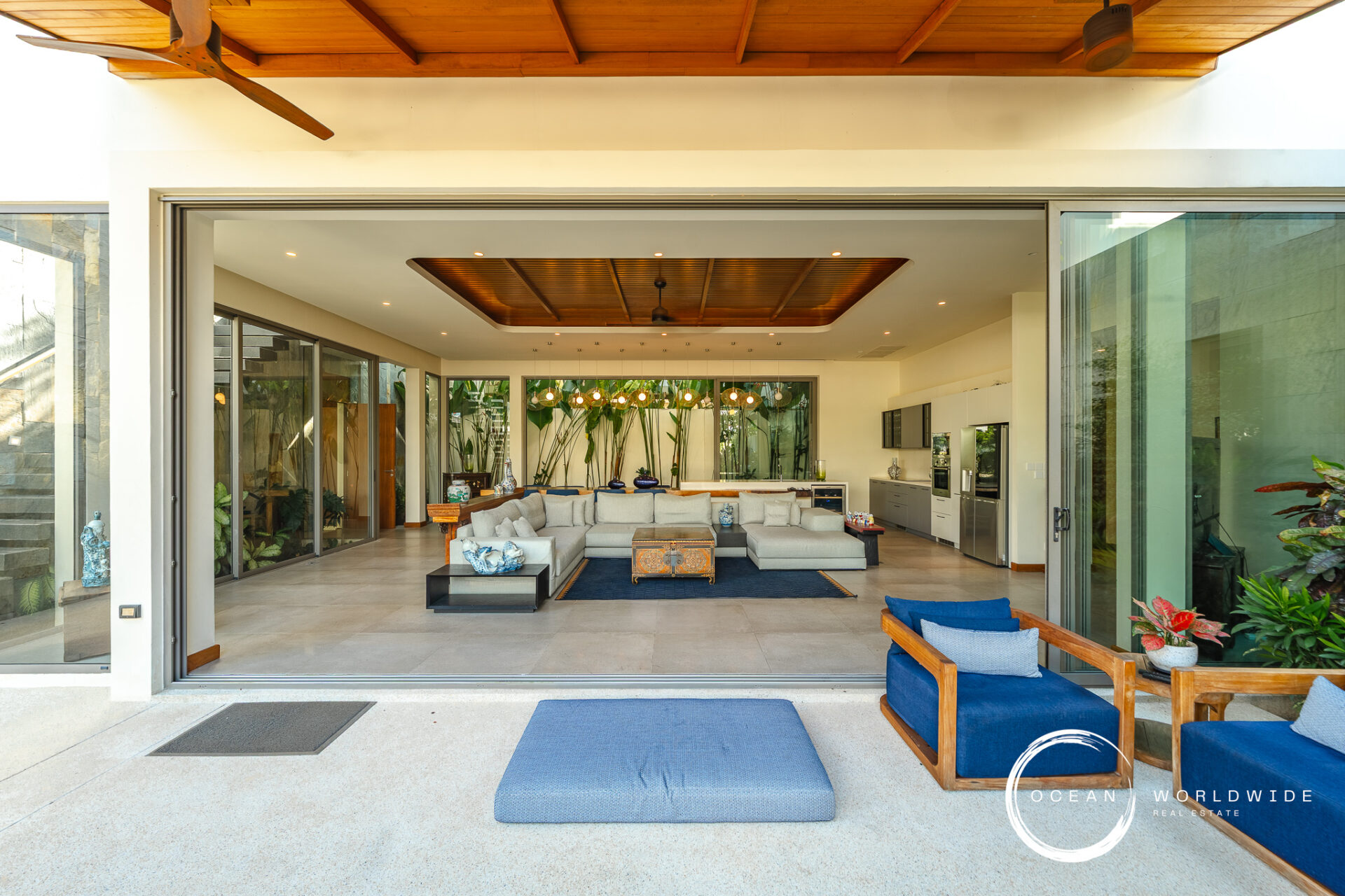 Open-plan living room seen through wide sliding doors; gray sectional sofa, blue cushions, tropical plants, and a kitchen in the background.