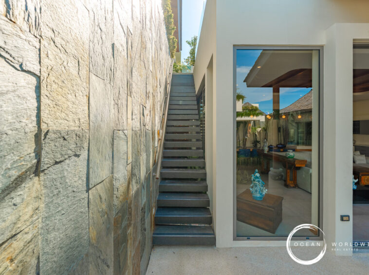 Narrow outdoor passage with a stone wall on the left and a modern glass-fronted house on the right, featuring a grey metal staircase between them.