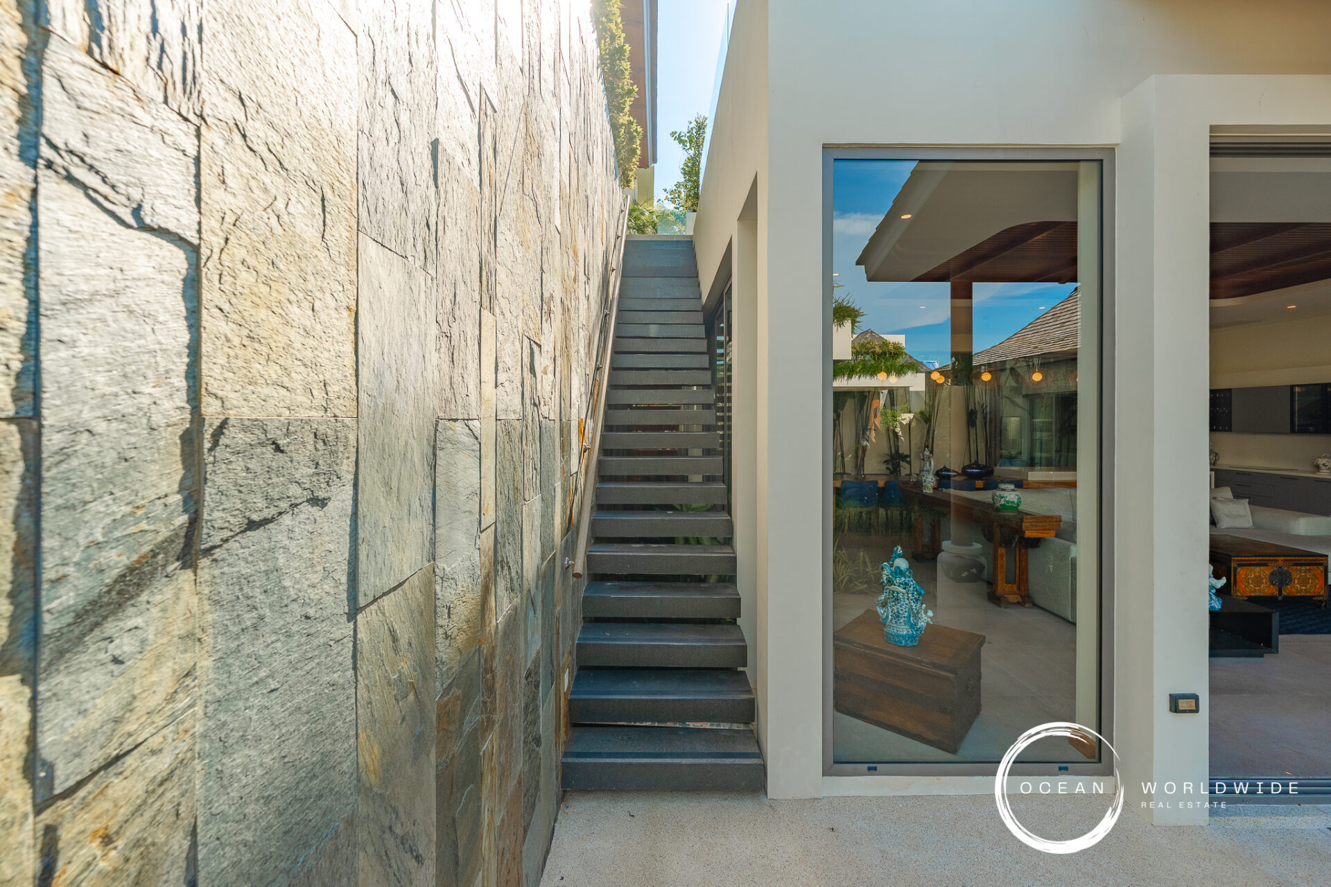 Narrow outdoor passage with a stone wall on the left and a modern glass-fronted house on the right, featuring a grey metal staircase between them.