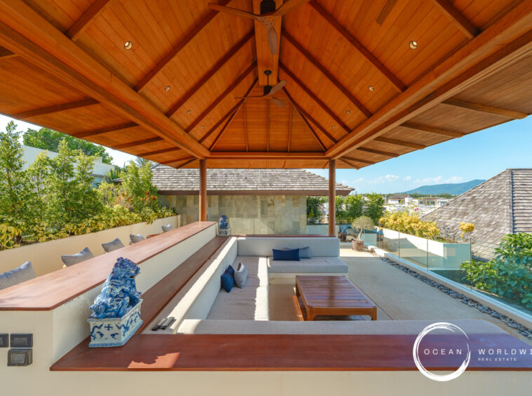 Outdoor lounge under a wooden vaulted ceiling with built-in beige seating, blue pillows, and glass railing overlooking rooftops and hills.