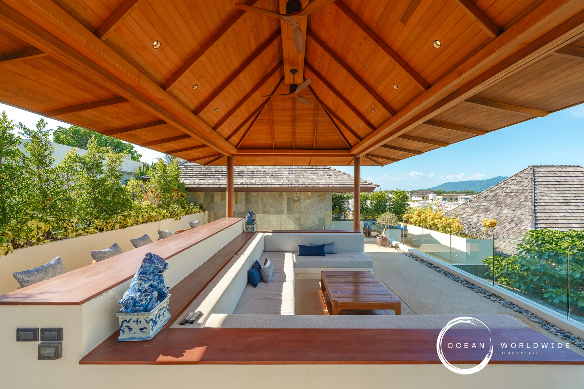 Outdoor lounge under a wooden vaulted ceiling with built-in beige seating, blue pillows, and glass railing overlooking rooftops and hills.