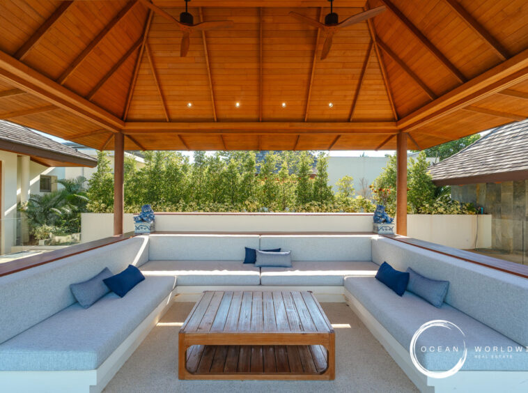 Outdoor covered seating area with a U-shaped light-gray sectional, blue pillows, and a wooden coffee table under a wooden pavilion. Clear garden and greenery beyond the seating area, plus an Ocean Worldwide Real Estate watermark in the corner.