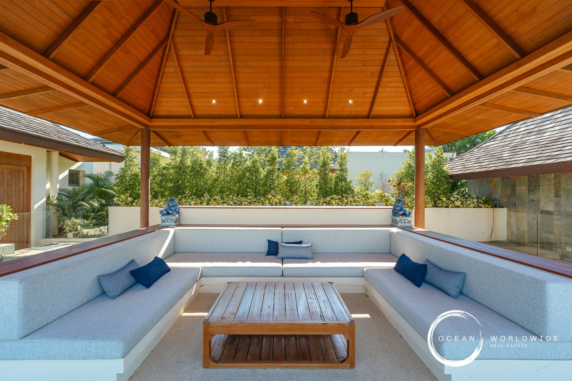 Outdoor covered seating area with a U-shaped light-gray sectional, blue pillows, and a wooden coffee table under a wooden pavilion. Clear garden and greenery beyond the seating area, plus an Ocean Worldwide Real Estate watermark in the corner.