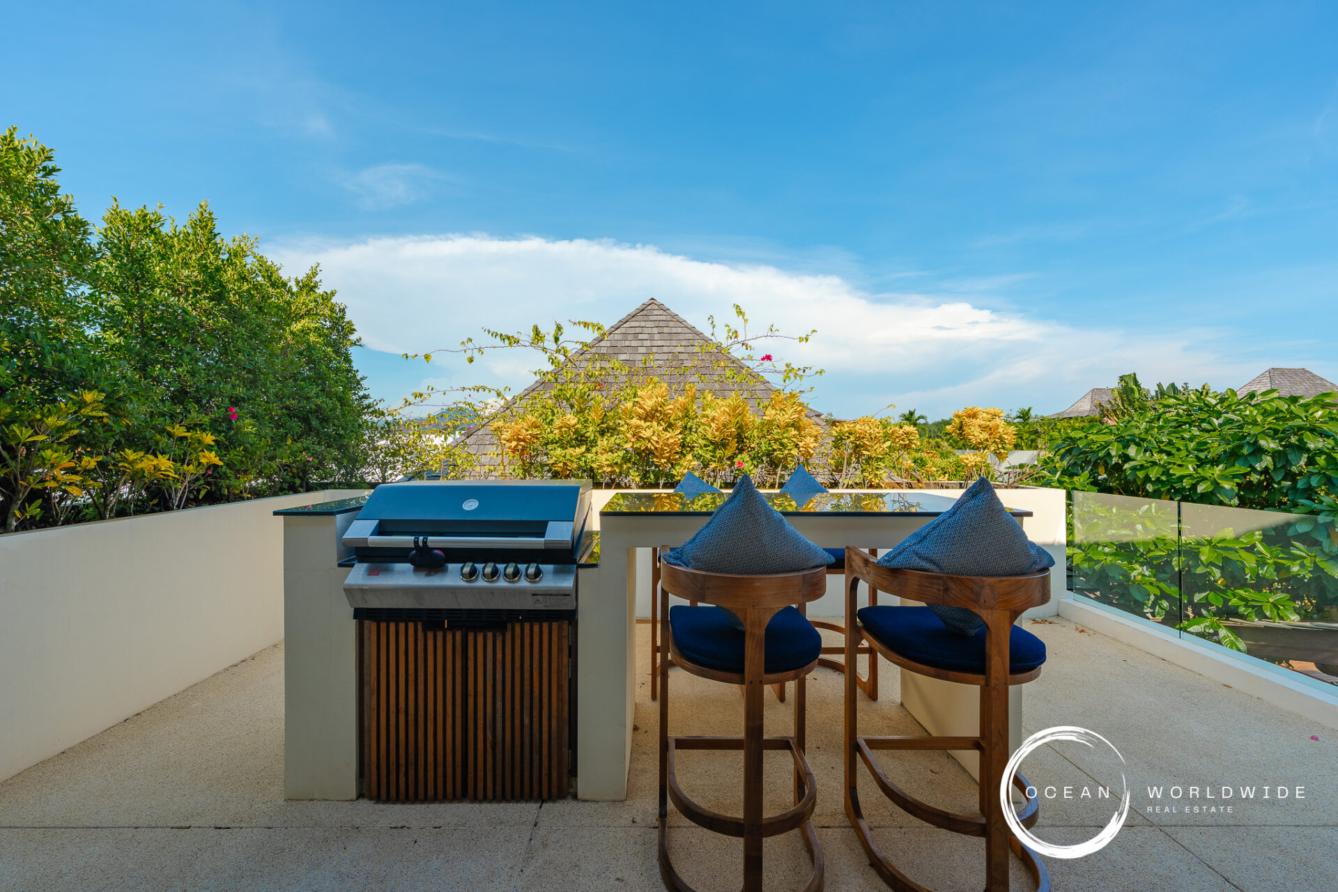 Outdoor balcony with a grill and two wooden chairs with blue cushions, glass railing, tropical plants, and a blue sky; Ocean Worldwide Real Estate watermark in corner.