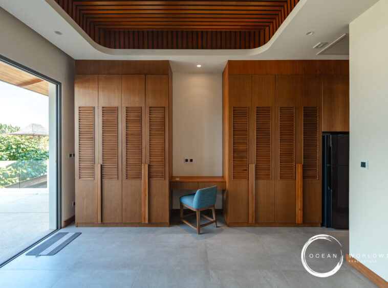 Interior with tall wooden wardrobes featuring louvered doors, a small desk and blue chair, and a wood slatted ceiling; glass sliding door reveals greenery outside.