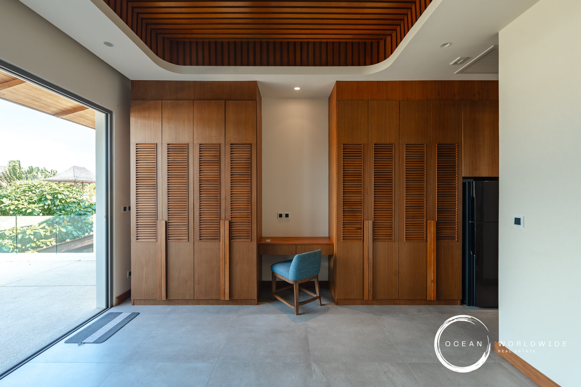 Interior with tall wooden wardrobes featuring louvered doors, a small desk and blue chair, and a wood slatted ceiling; glass sliding door reveals greenery outside.