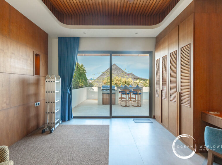 Indoor hallway with warm wooden wall panels leading to a glass balcony door; outside, a patio with bar stools and a grill against a bright blue sky. A blue curtain hangs to the left. Ocean Worldwide watermark in corner.