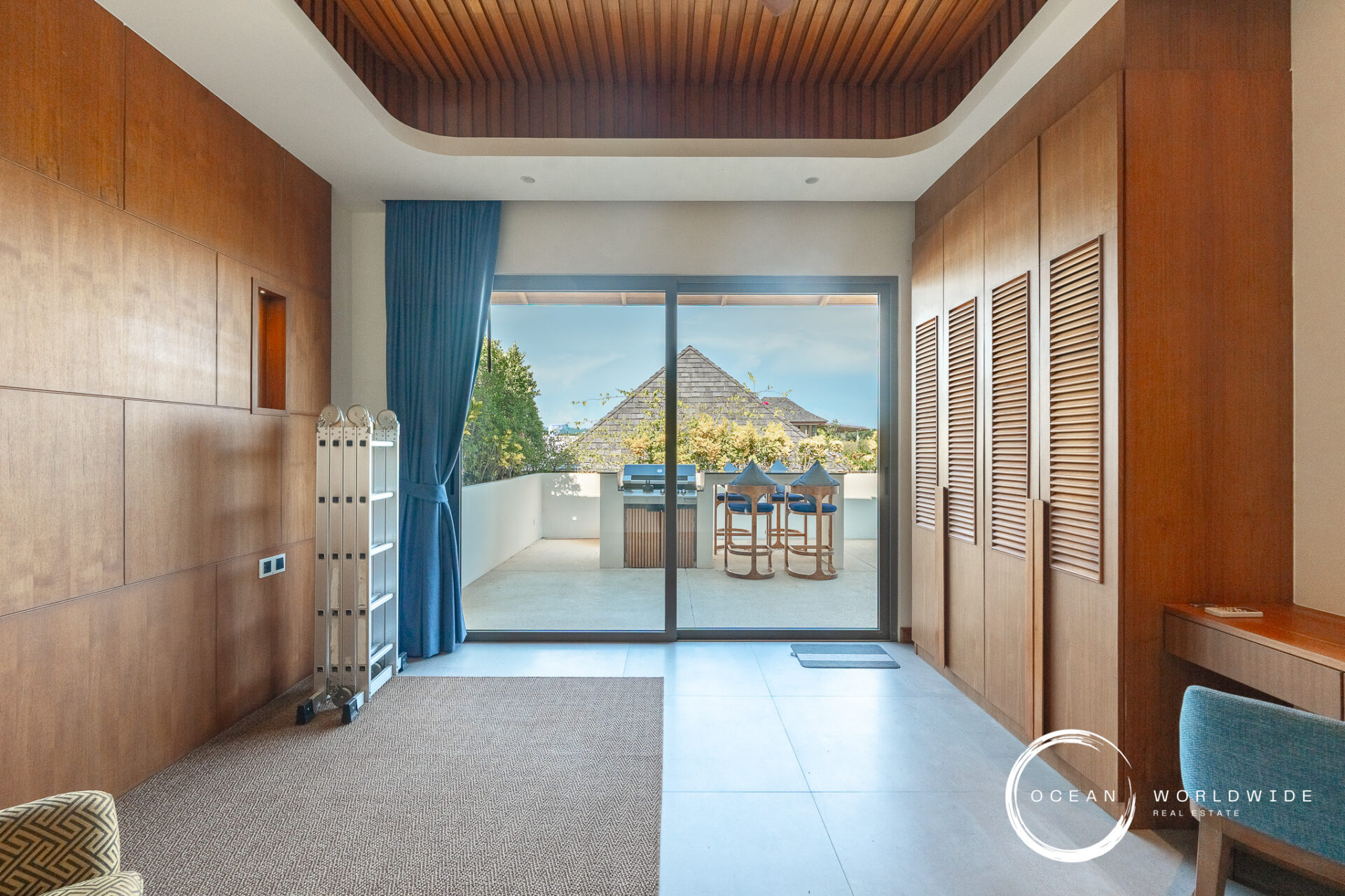 Indoor hallway with warm wooden wall panels leading to a glass balcony door; outside, a patio with bar stools and a grill against a bright blue sky. A blue curtain hangs to the left. Ocean Worldwide watermark in corner.