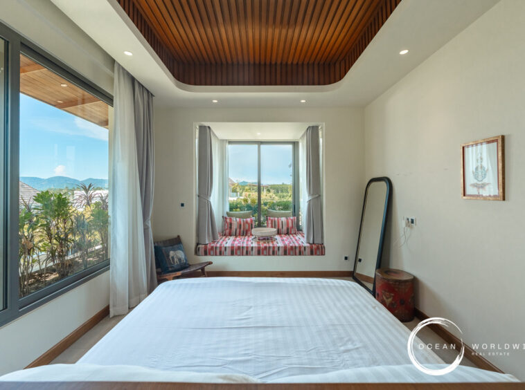 Bright hotel bedroom with a large window seating nook featuring red plaid cushions, white curtains, and a view of greenery and hills; wood-paneled ceiling above and white walls with a framed artwork.
