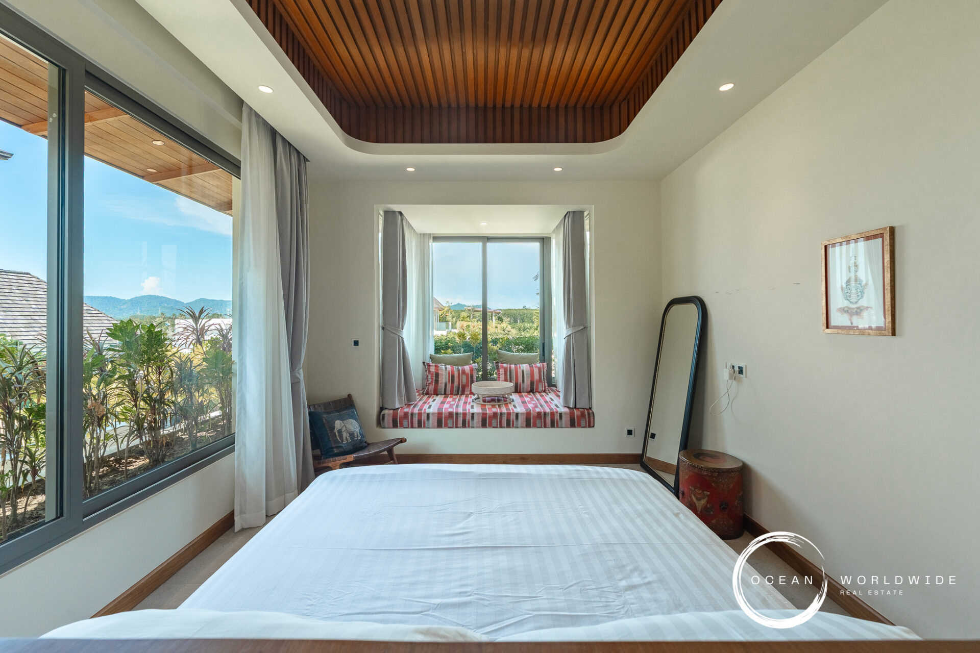 Bright hotel bedroom with a large window seating nook featuring red plaid cushions, white curtains, and a view of greenery and hills; wood-paneled ceiling above and white walls with a framed artwork.