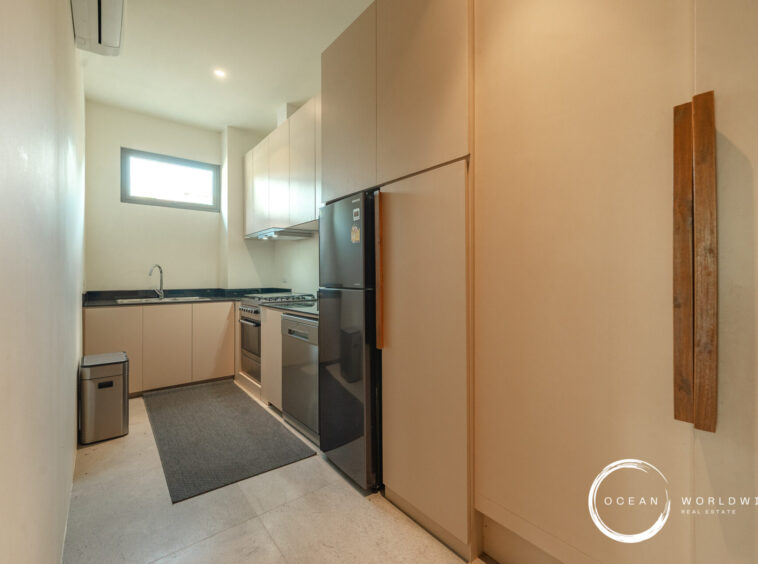 Narrow modern galley kitchen with beige cabinetry, black countertops, a stainless steel fridge and stove, and a small horizontal window above the sink.