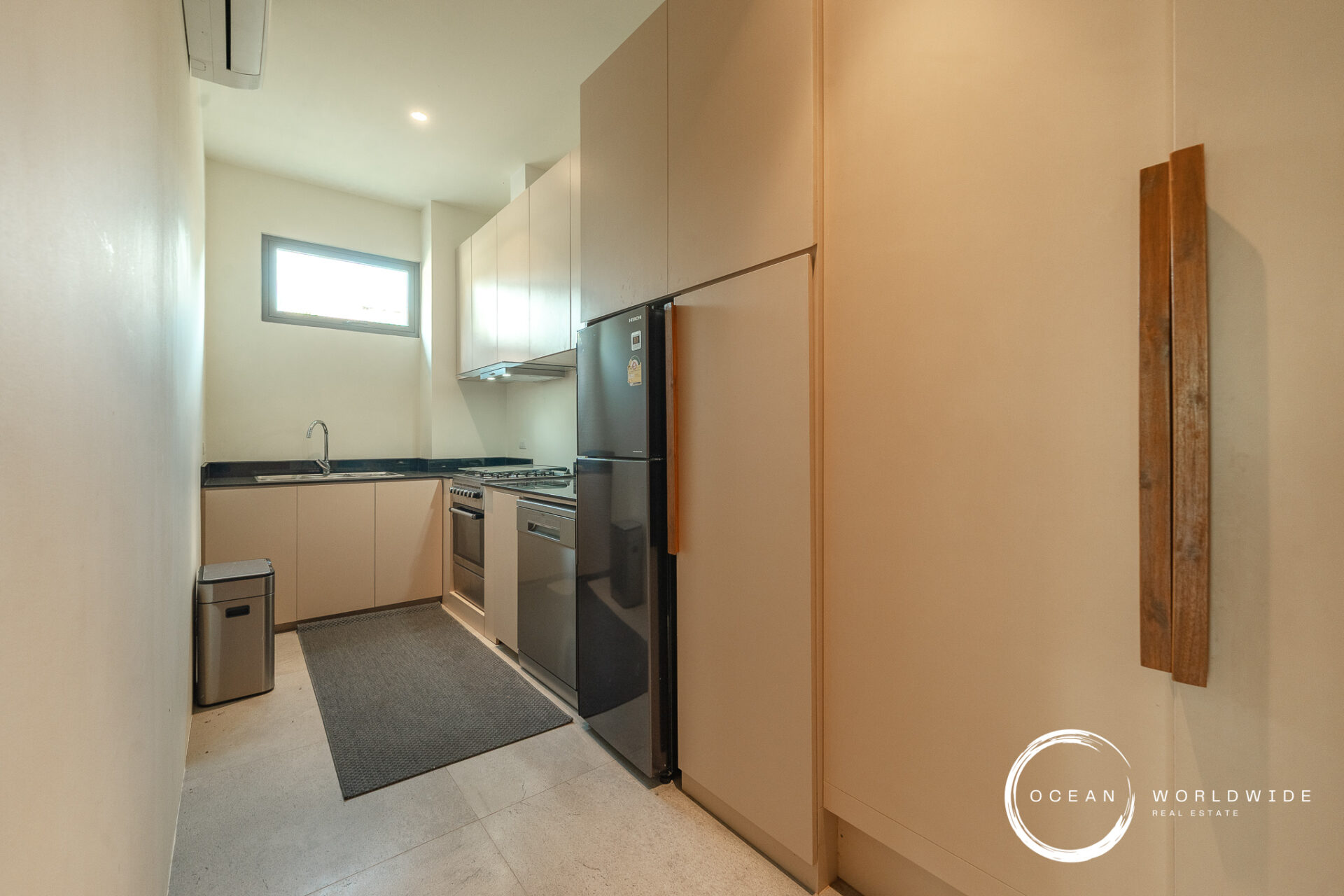 Narrow modern galley kitchen with beige cabinetry, black countertops, a stainless steel fridge and stove, and a small horizontal window above the sink.