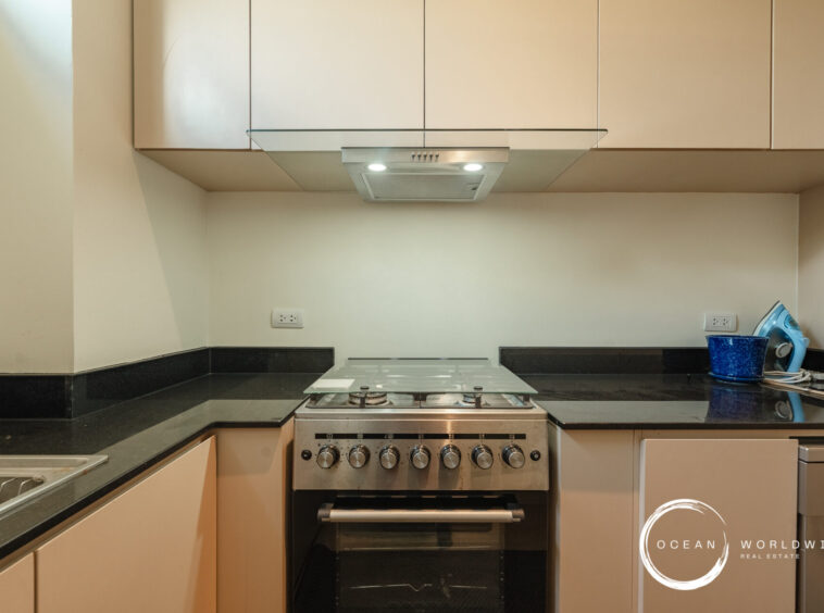 Modern kitchen with a stainless steel gas range centered under a glass canopy hood, black granite countertops, and beige upper cabinets; sink on the left and an iron on the right with a blue mug on the counter; watermark in bottom-right corner reading 'Ocean Worldwide Real Estate'.