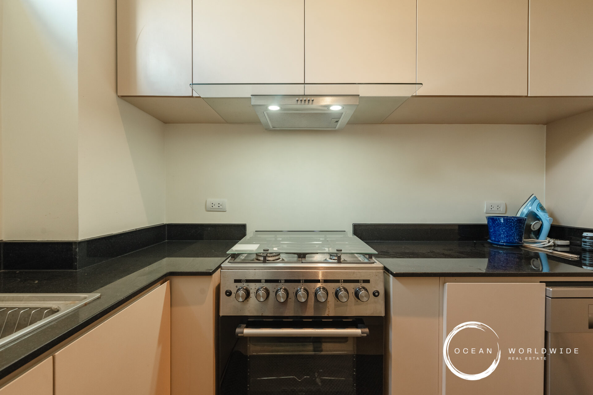 Modern kitchen with a stainless steel gas range centered under a glass canopy hood, black granite countertops, and beige upper cabinets; sink on the left and an iron on the right with a blue mug on the counter; watermark in bottom-right corner reading 'Ocean Worldwide Real Estate'.