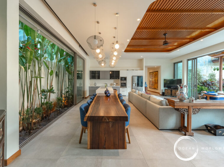 Open-plan living and dining area with a long solid-wood table, blue chairs, pendant globe lights, and a modern kitchen in the background.