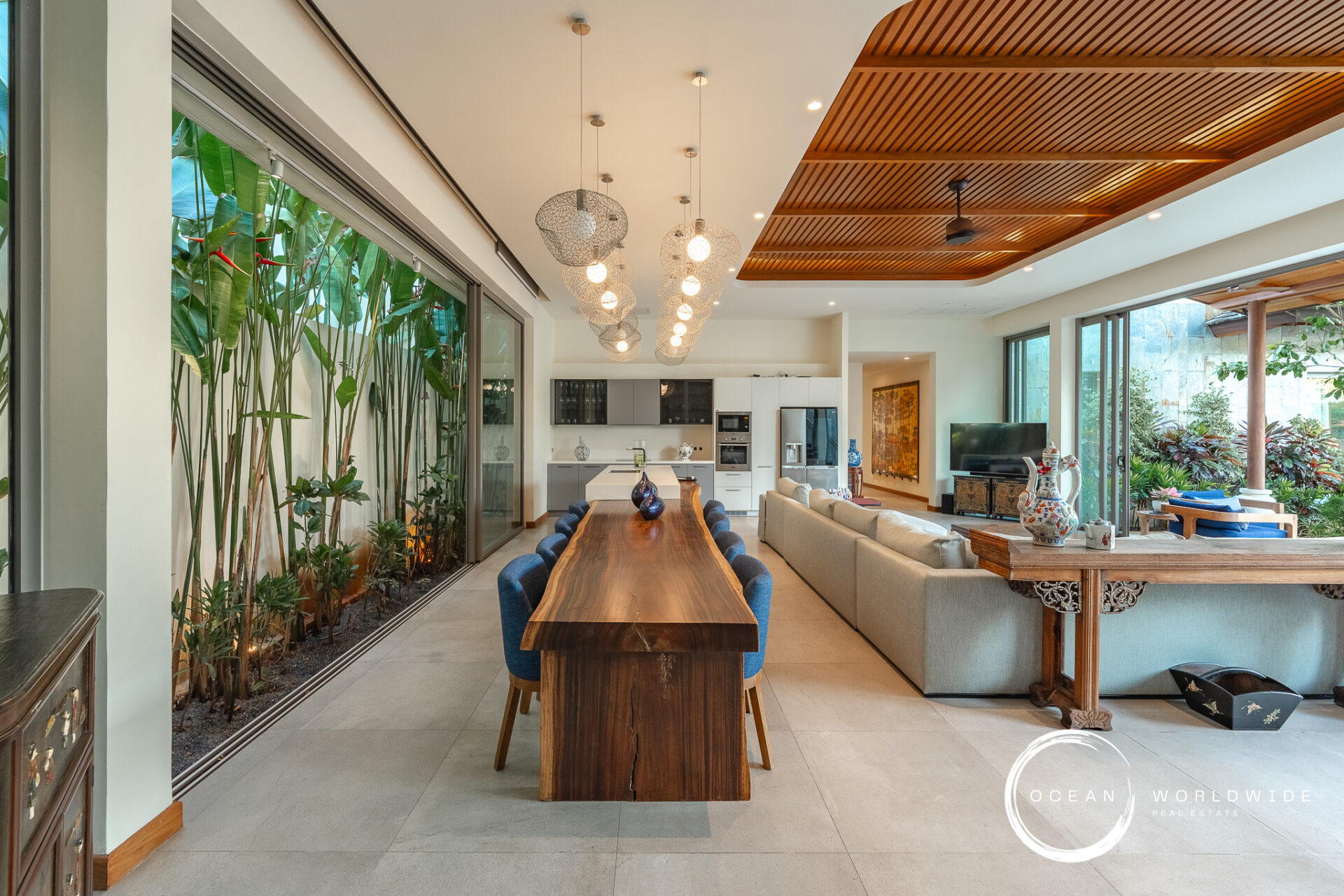 Open-plan living and dining area with a long solid-wood table, blue chairs, pendant globe lights, and a modern kitchen in the background.