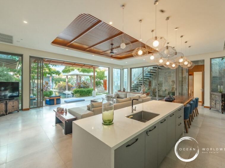 Open-plan living area with a large white kitchen island, gray cabinetry, and a row of blue bar stools facing a spacious living room and outdoor garden view.