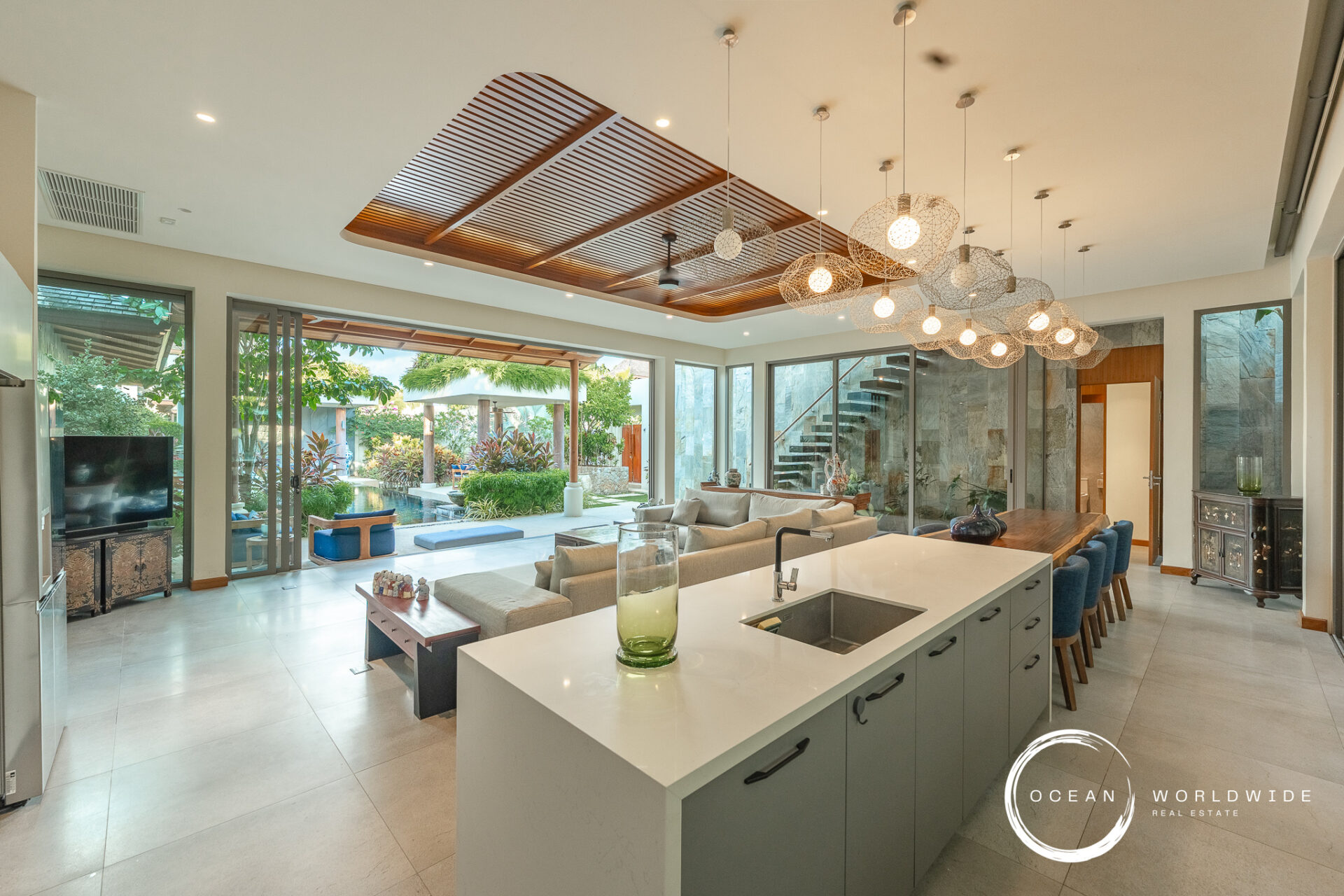 Open-plan living area with a large white kitchen island, gray cabinetry, and a row of blue bar stools facing a spacious living room and outdoor garden view.
