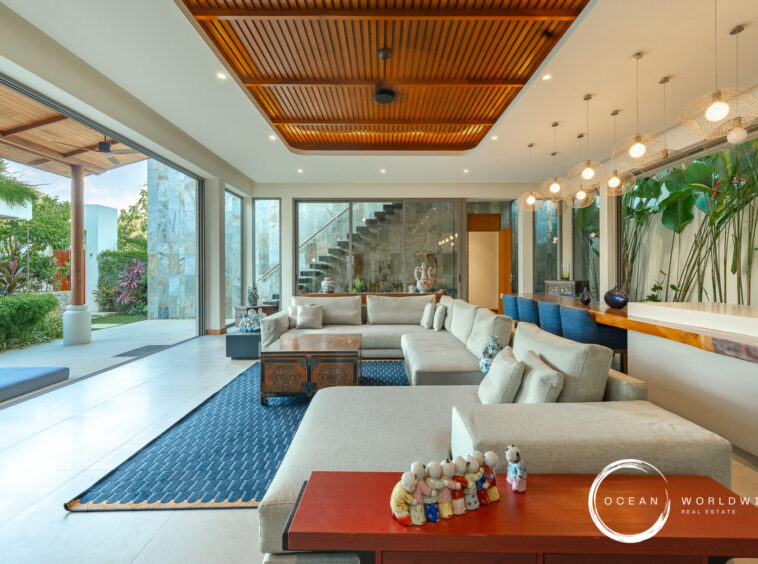 Spacious modern living room with beige sofas, glass doors to a garden, and a wooden slat ceiling. Outdoor greenery visible outside.