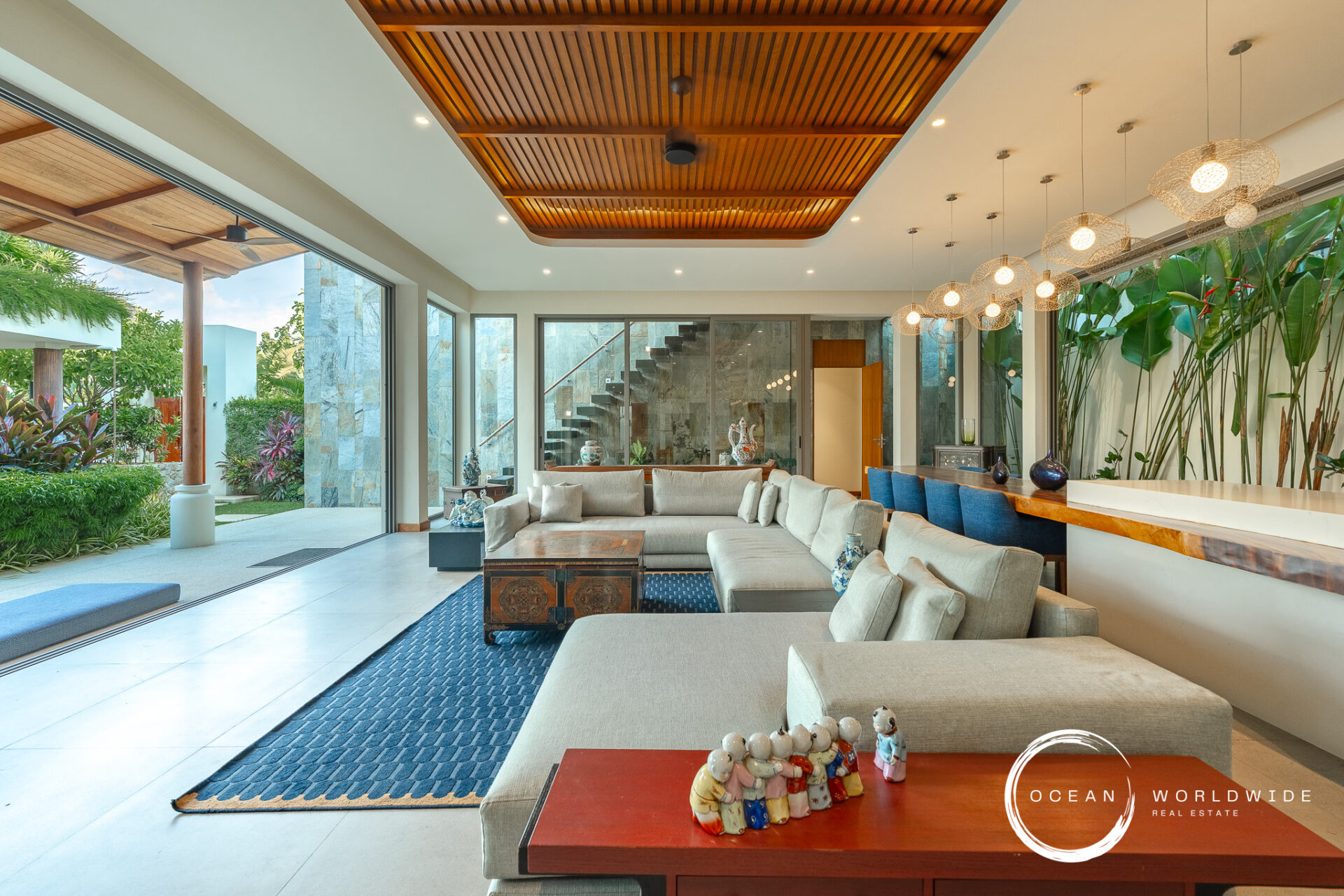 Spacious modern living room with beige sofas, glass doors to a garden, and a wooden slat ceiling. Outdoor greenery visible outside.