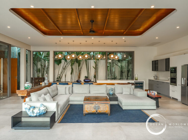 Open living area with a gray sectional, antique chest coffee table, and a blue rug beneath a wood-paneled ceiling and pendant lights in a bright kitchen.