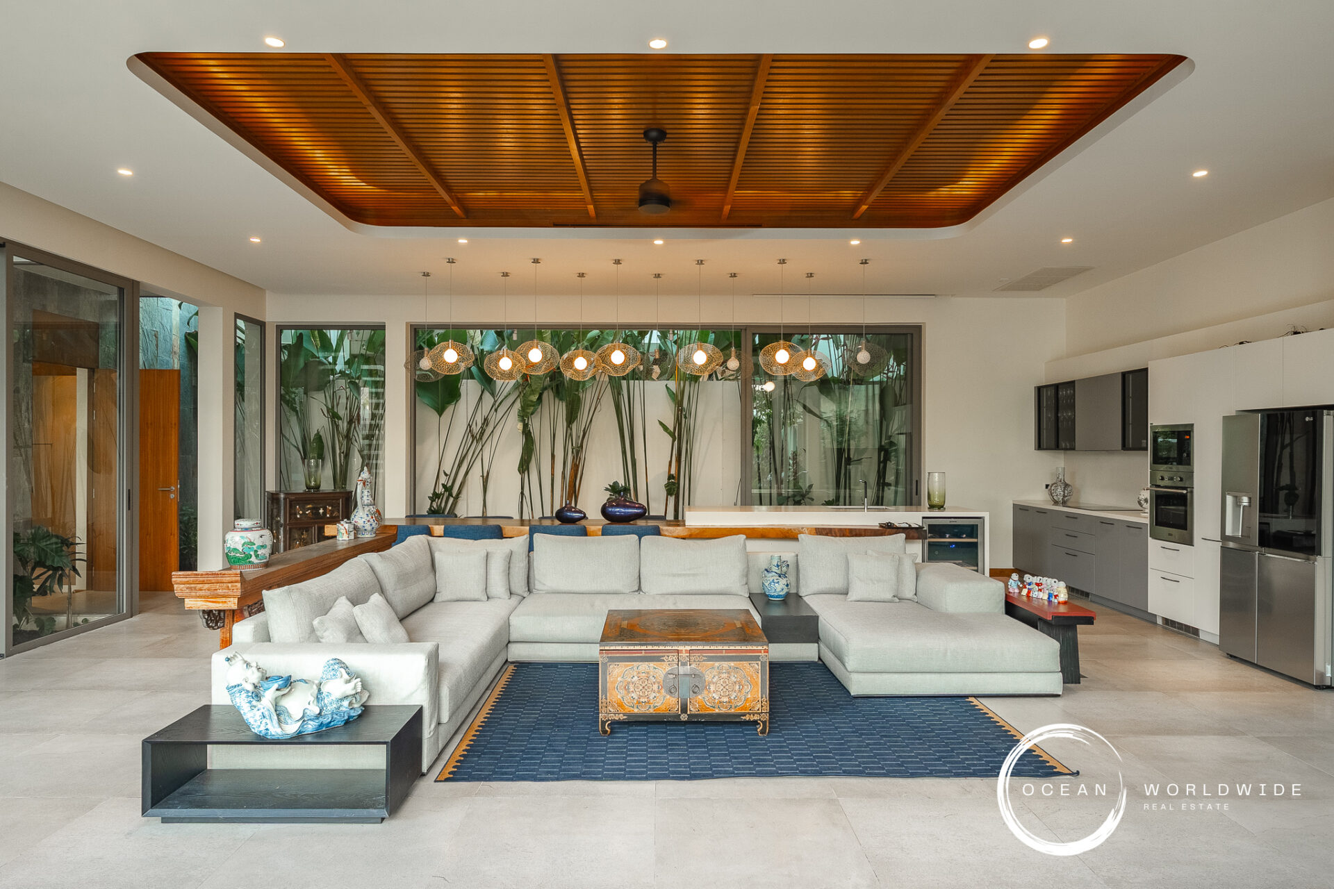 Open living area with a gray sectional, antique chest coffee table, and a blue rug beneath a wood-paneled ceiling and pendant lights in a bright kitchen.