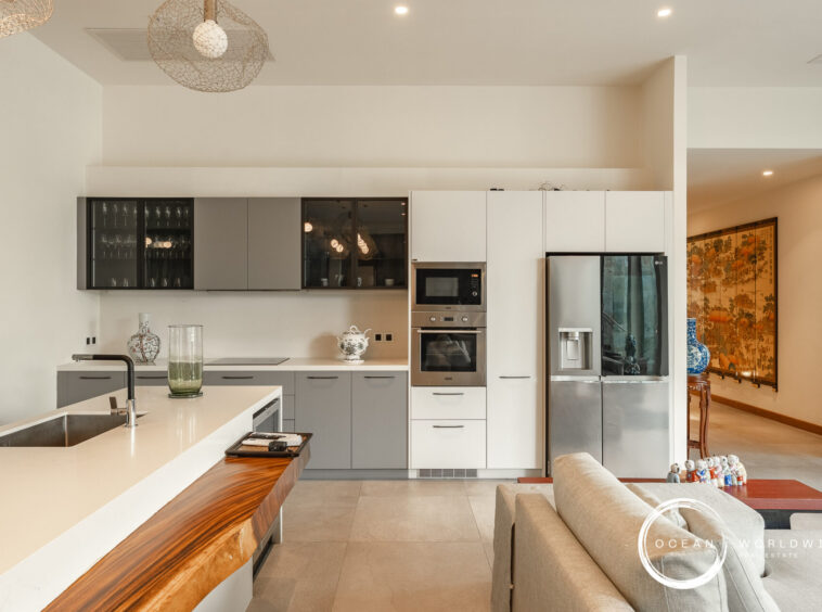 Modern open-plan kitchen with a white island, gray lower cabinets, and stainless steel refrigerator and double ovens; decorative vases on the counter and glass-front upper cabinets. The living area with a wooden screen and Asian artwork is visible through a doorway.