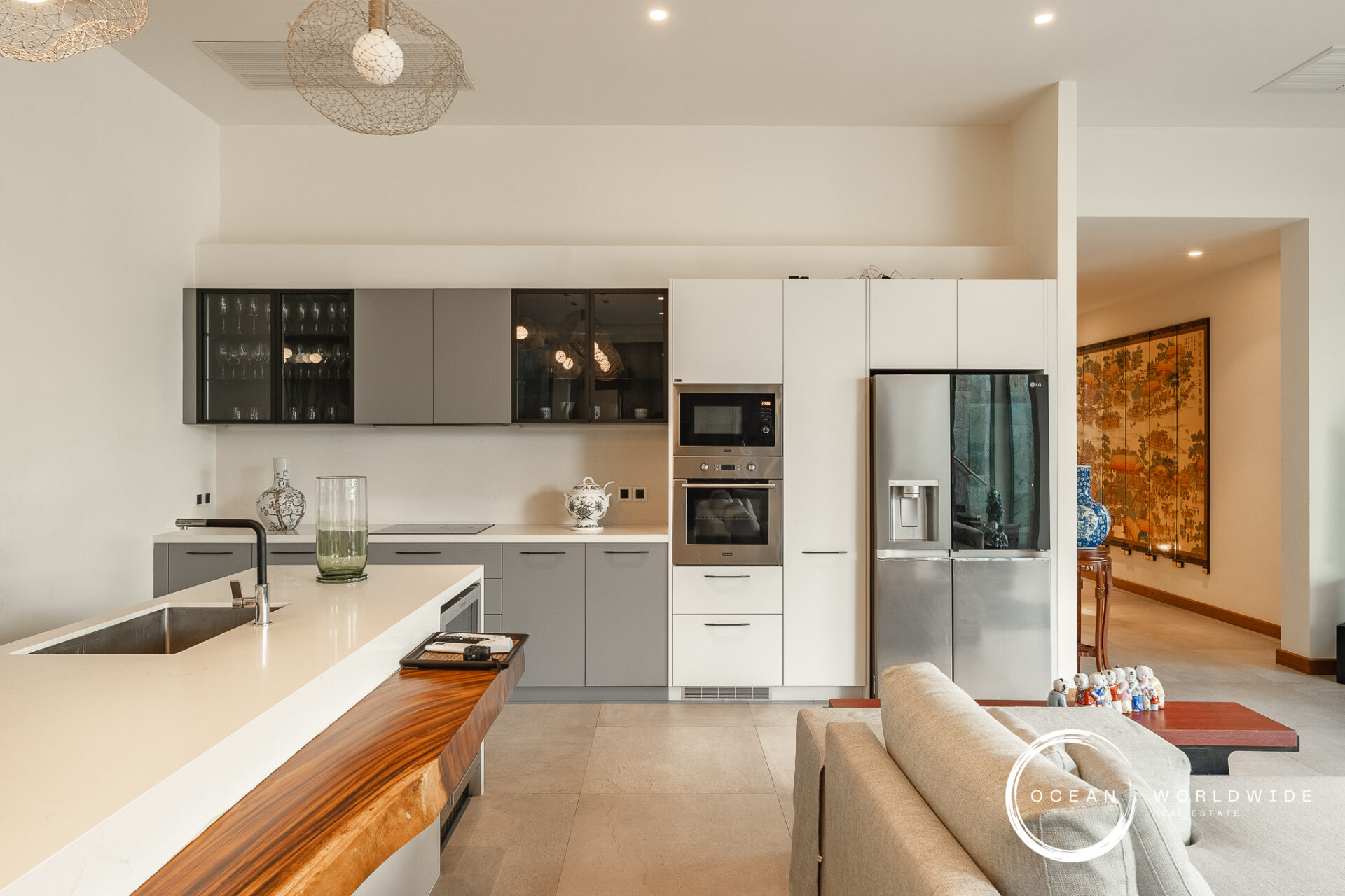 Modern open-plan kitchen with a white island, gray lower cabinets, and stainless steel refrigerator and double ovens; decorative vases on the counter and glass-front upper cabinets. The living area with a wooden screen and Asian artwork is visible through a doorway.